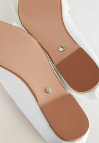 Chaussures en satin blanc avec une finition lisse, dotées d'une semelle en caoutchouc beige clair et de détails métalliques sur le talon. Design plat et arrondi.