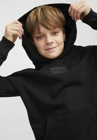 Sudadera negra con bolsillo frontal, que presenta un parche de marca texturizado. La tela parece suave y flexible, con un diseño ajustado y capucha ajustable.