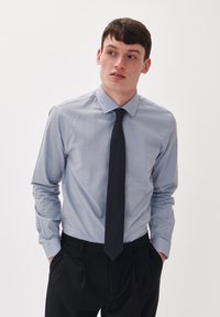 Lichtblauw overhemdd met een klein stippenpatroon, met knoopsluiting op de kraag, lange mouwen en een donkere stropdas, gedragen met zwarte broek.