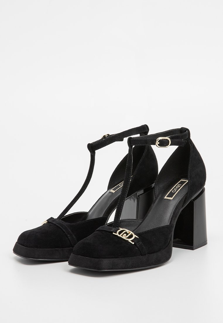 Tacones altos de ante negro con tacón cuadrado, diseño de tirantes en T, punta cerrada y acento de logo dorado en el frente. Superficie texturizada.