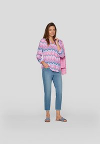 Leichte Bluse mit rosa und blauen Zickzackmustern und langen Ärmeln, kombiniert mit blauen, cropped Jeans und grauen Sandalen.