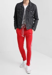 Černá džínová bunda, bílý tričko, červené sportovní kalhoty s bílými bočními pruhy a bílé tenisky. Model stojí na neutrálním pozadí.
