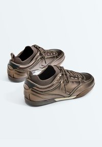Paire de baskets à lacets bronze métallisé avec col en maille autour de la cheville et accents noirs au talon, présentée sur fond blanc.