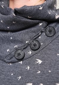 Tissu gris à la texture douce orné de motifs d'oiseaux blancs. Trois boutons noirs servent de fermoirs sur un col bénitier.