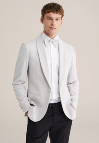 Tânărul poartă o jachetă de smoking gri deschis, cămașă albă, papion asortat și pantaloni negri, stând cu mâinile în buzunare.