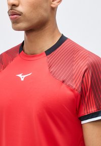 Άνδρας που φοράει κόκκινο αθλητικό πουκάμισο με μαύρο στρογγυλό λαιμό και ριγέ μανίκια, με λευκό λογότυπο Mizuno στο στήθος.