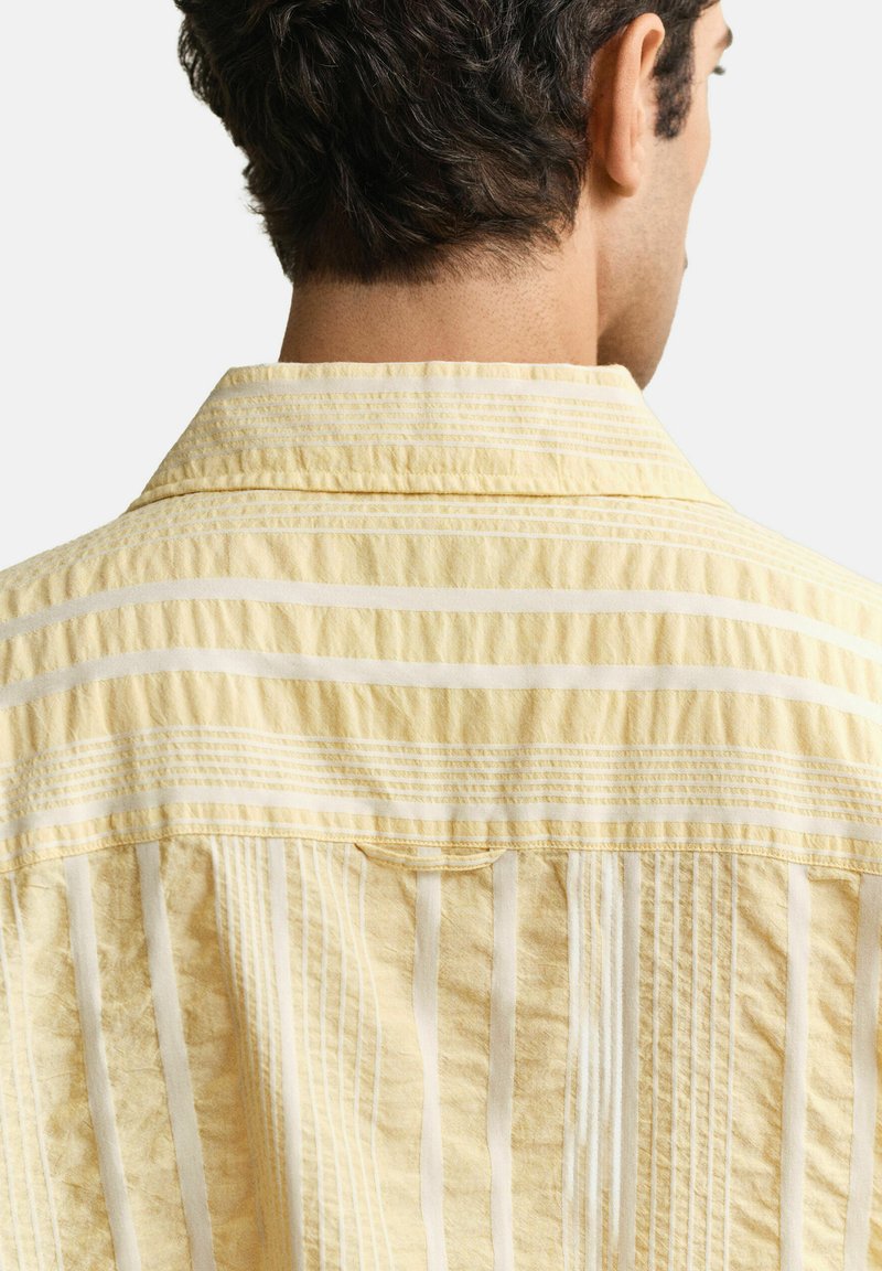 Mann von hinten zu sehen, trägt ein gelb-weiß gestreiftes Hemd mit vertikalen Streifen und einem umgeklappten Kragen vor einfachem Hintergrund.