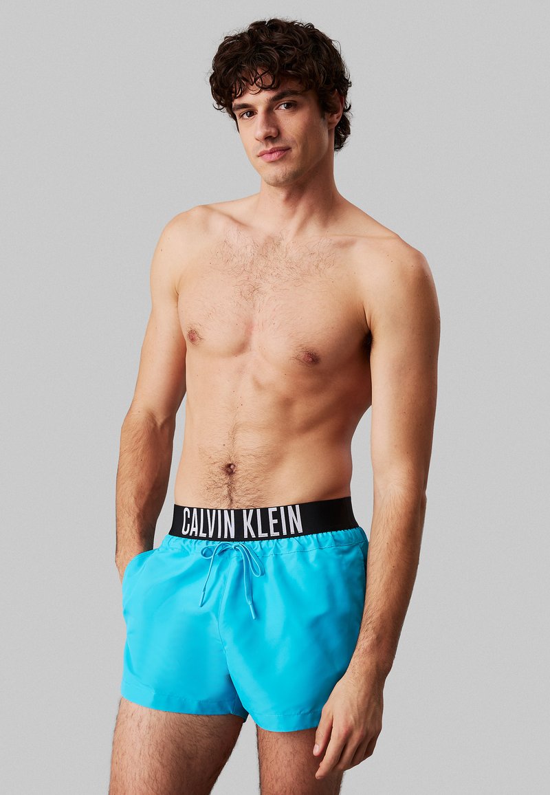 Costumi da bagno turchesi realizzati in tessuto leggero, con una cintura elastica nera di Calvin Klein e un cordoncino per la regolazione.