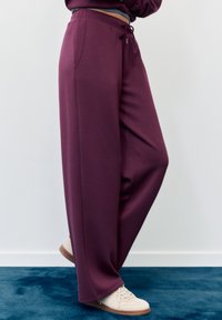 Bordowe spodnie o szerokich nogawkach wykonane z miękkiej tkaniny, z elastycznym pasem i sznurkiem oraz kieszeniami po bokach, połączone z beżowymi sneakersami.