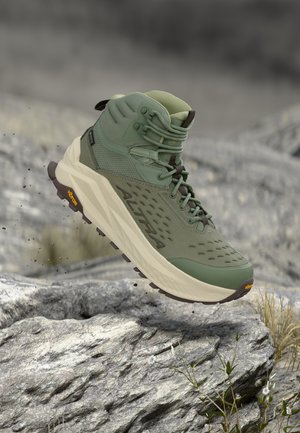 Bocanc verde înalt pentru drumeții, cu talpă groasă bej, talpă exterioară Vibram și branding Altra, plutind deasupra unui teren stâncos cu plante mici.