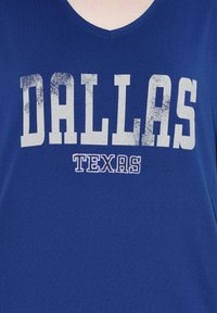 Modrá košile s výstřihem do V, na které je velkým bílým opotřebovaným písmem nápis "DALLAS" a pod ním menším obrysovým fialovým písmem nápis "TEXAS".