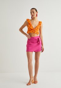Top corto floral naranja con mangas acampanadas, combinado con una falda envolvente rosa con un detalle de lazo. Llevado con sandalias de tacón naranjas.