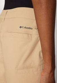 Calções beges feitos de tecido leve, com um bolso traseiro e o logo da Columbia em preto. Textura suave com detalhes de costura subtil.