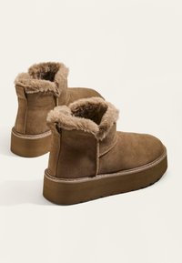 Stivali ankle in suede marrone con bordo di pelliccia, dotati di una suola platform spessa e semplici dettagli di cucitura. L'interno morbido è visibile.