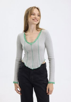 Gray ribbed long-sleeve top with green ruffle accents along the neckline and hem, paired with black high-waisted trousers.