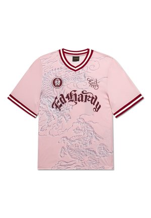 Junger Mann mit dunklen Haaren trägt ein pinkes Grafik-T-Shirt, hellblaue Jeansshorts, weiße Socken und beige Sneakers, steht vor einem schlichten grauen Hintergrund.