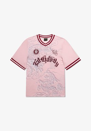 Junger Mann mit dunklen Haaren trägt ein pinkes Grafik-T-Shirt, hellblaue Jeansshorts, weiße Socken und beige Sneakers, steht vor einem schlichten grauen Hintergrund.