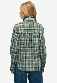 Blonde Frau, die sich abwendet, trägt ein langärmliges Hemd in grün-grauem Karomuster mit hohem Kragen und blaue Jeans.