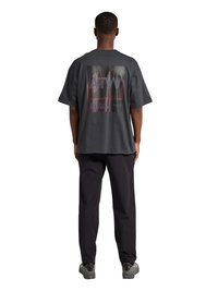 Mężczyzna stoi tyłem, ubrany w luźną ciemnoszarą koszulę z delikatnym nadrukiem na plecach, czarne spodnie i szare trampki.