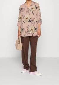 Långärmad, ljusrosa blus med blommönster, i kombination med bruna, löst sittande byxor och vita sneakers med rosa detaljer. Håller en vävd handväska.