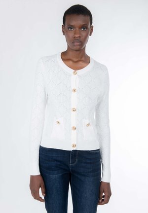 Weiß gestrickter Cardigan mit Rautenmuster, 7 goldenen Knöpfen und zwei aufgesetzten Taschen vorne. Abgeschlossen mit einem abgerundeten Ausschnitt und gerippten Bündchen.
