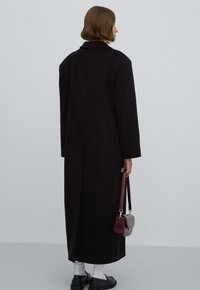 Lange zwarte jas met een gestructureerd ontwerp, brede mouwen en een reverskraag. Gecombineerd met een kleine bordeauxrode handtas en witte sokken.