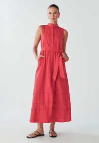 Šaty do dĺžky kolien, bez rukávov, v žiarivej koralovej farbe. Obsahujú zapínanie na gombíky, zúžený pás s rovnakým opaskom a bočné vrecká. Hladká textura.