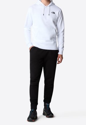 Vit hoodie med framficka och svart logga, kombinerad med svarta joggers och sportskor i flera färger.