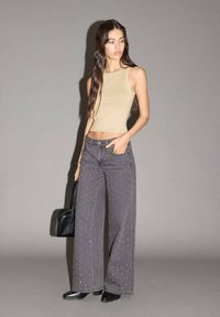 Žena s dlouhými tmavými vlasy, oblečená v béžovém bezrukávovém crop topu, šedých širokých kalhotách se cvočky, černých botách a držící černou kabelku.