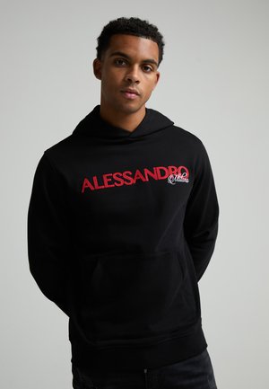 Jeune homme aux cheveux courts et bouclés portant un sweat à capuche noir avec "ALESSANDRO Milano" écrit en rouge et blanc sur la poitrine.