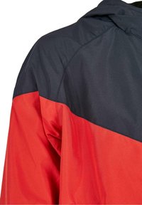 Veste color-block rouge et marine, fabriquée en tissu lisse et léger. Dotée d'une capuche et de détails de couture visibles.