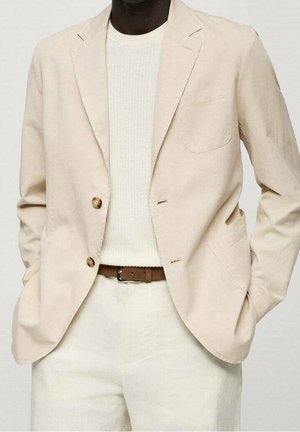 Mężczyzna w beżowej marynarce na białym swetrze dzianym i kremowych spodniach z brązowym paskiem, ręce w kieszeniach marynarki.