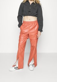 Avkortad svart zip-up sweatshirt, orange fusk läderbyxor med sidoklyftor och tryckknappdetaljer, kombinerat med vita sneakers med svarta ränder.