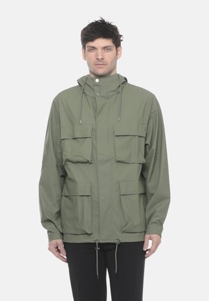 Uomo che indossa giacca con cappuccio verde oliva con quattro tasche anteriori con patta e coulisse, in piedi su sfondo bianco.