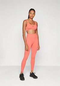 Conjunto deportivo color durazno que incluye un sujetador deportivo recortado y leggings de cintura alta, hechos de un material elástico. Zapatillas negras completan el conjunto.