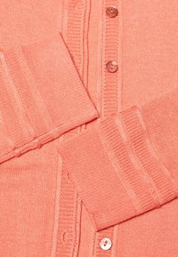 Korálový pletený cardigan s žebrovanými manžetami, pěti knoflíky a texturovaným vzorem. Materiál je měkký a lehký.