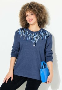 Sweatshirt bleu marine avec des embellissements en sequins de différentes nuances de bleu, associé à un petit sac bleu et un pantalon noir.