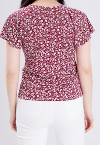 Blouse florale rouge avec des manches courtes évasées, présentant un dos froncé et un motif floral blanc sur un tissu texturé.