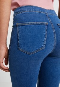 Person wearing high-waisted blue denim jeans with back pockets and a tucked-in pink shirt, shown from the waist down.