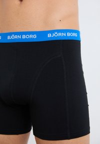Schwarze Boxershorts aus glatt gewebtem Stoff mit einem blauen Gummibund, der das Logo "BJÖRN BORG" in weißer Schrift trägt.