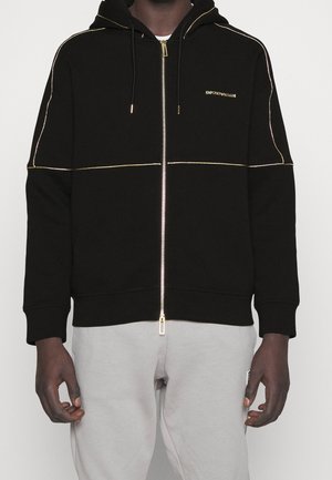 Musta vetoketjuhuppari, jossa kultaiset somisteet ja Emporio Armani -logo, päällä henkilö, jolla on vaaleanharmaat housut ja yksivärinen tausta.