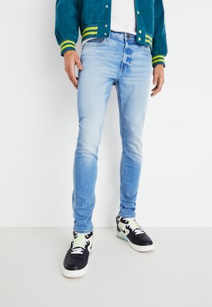 Jeans slim-fit azzurri, con effetto sbiadito. Indossati con sneakers nere con accenti verdi e una giacca in tessuto texture.