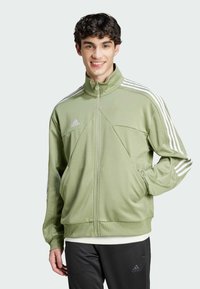 Giacca zip-up verde chiaro con colletto alto, logo Adidas bianco e tre strisce bianche lungo le maniche. Tessuto liscio e vestibilità rilassata.