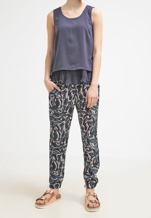 Vrouw die een mouwloze marineblauwe top draagt, joggingbroek met varenprint en metallic roze-gouden sandalen met gespen, staand met handen in de zakken.