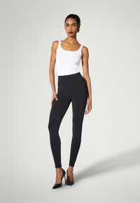 Leggings nere con vita alta e vestibilità elasticizzata, abbinate a un top bianco aderente e décolleté nere con punta affilata. Texture liscia, design minimalista.
