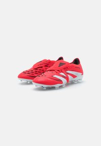 Scarpe da football rosse con tomaia testurizzata, tre strisce bianche prominenti sul lato, fissate con una fascia in Velcro e suola trasparente con tacchetti.