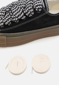 Zapatilla negra con gruesos cordones de patrón blanco y negro y dos cordones blancos enrollados sobre un fondo blanco.