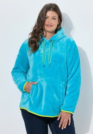 Donna che indossa una felpa con cappuccio in pile azzurro brillante con coulisse e rifiniture giallo neon, in piedi davanti a uno sfondo bianco con una mano nella tasca.