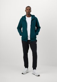 Teal zip-up hoodie with a front pocket, white t-shirt underneath, paired with black sweatpants and gray athletic shoes.