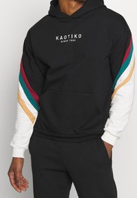 Felpa nera con la scritta "KAOTIKO Since 1999", strisce multicolore sulle maniche bianche, indossata con pantaloni neri, mano in tasca.